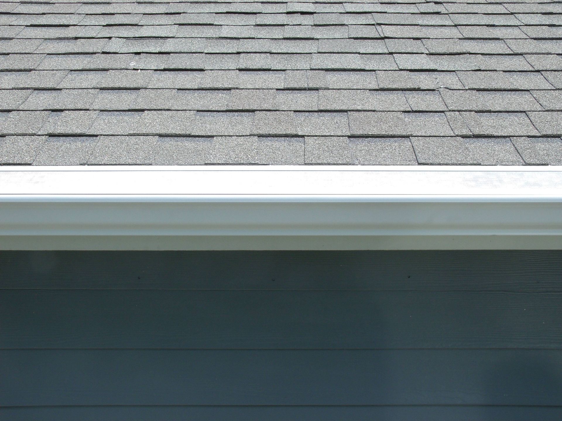 White gutter under gray shingle roof and blue siding of a house.
