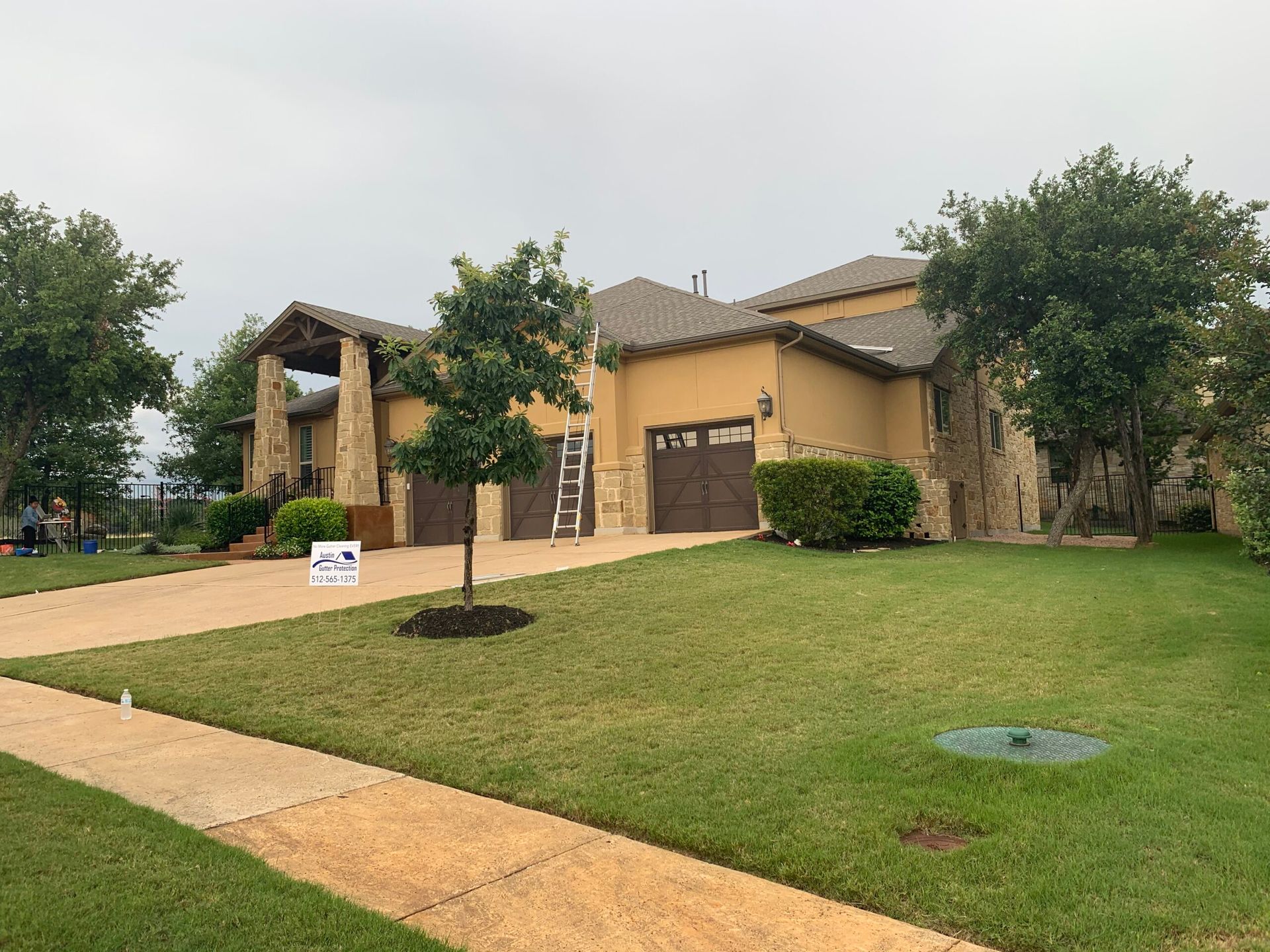 Two-story beige stucco house with brown roof, stone accents, two garage doors, and a green lawn.
