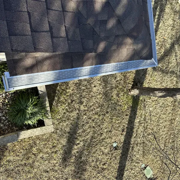Overhead view of a roof with shingles, gutter, and a dry, brown yard with a small plant.