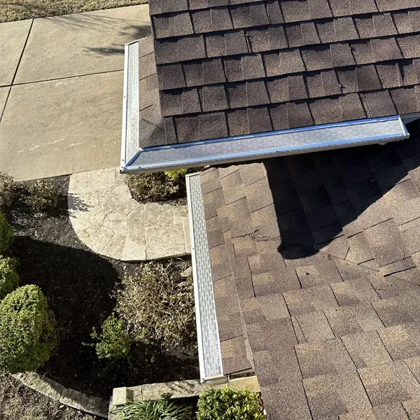 Overhead view of a house roof with brown shingles and a silver gutter, next to a concrete path and garden.