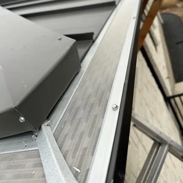 Gutter with metal mesh cover, bolted to the roof. Grey metal with a white brick wall in the background.
