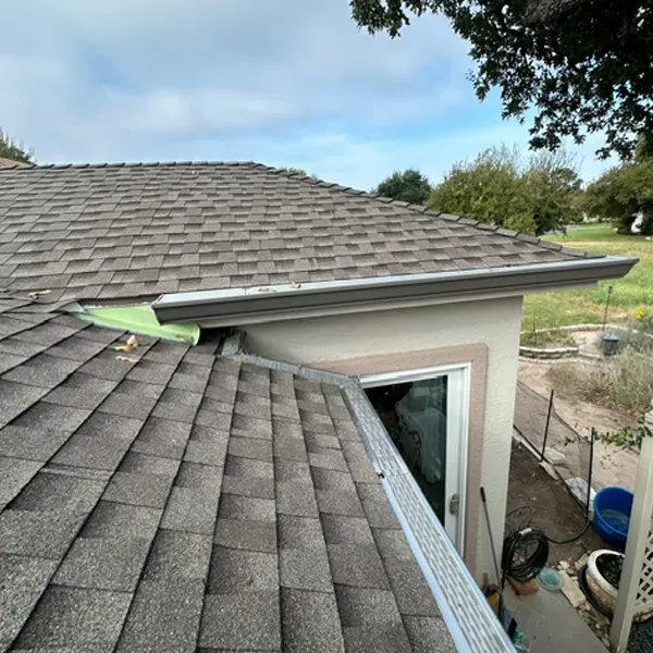 Overhead view of a roof with brown shingles, gray gutters, and a glimpse of a window and yard.