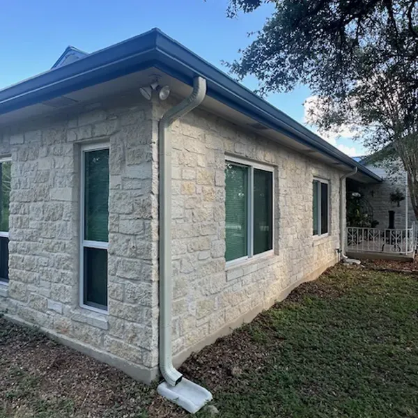 Beige stone building with white-framed windows, dark roof and gutters; grass and trees in background.