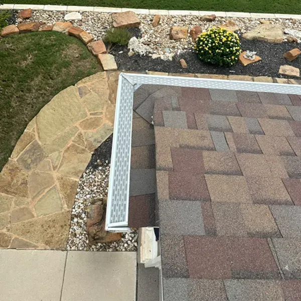 Overhead view of a roof with brown and tan shingles, a white gutter, and a stone walkway in a yard with landscaping.