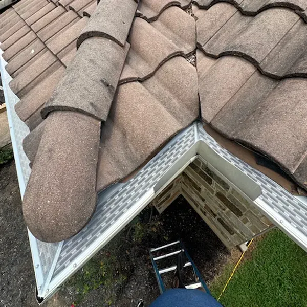 Close-up of a tiled roof with guttering and a ladder below. The tiles are brown, and the gutters are white.