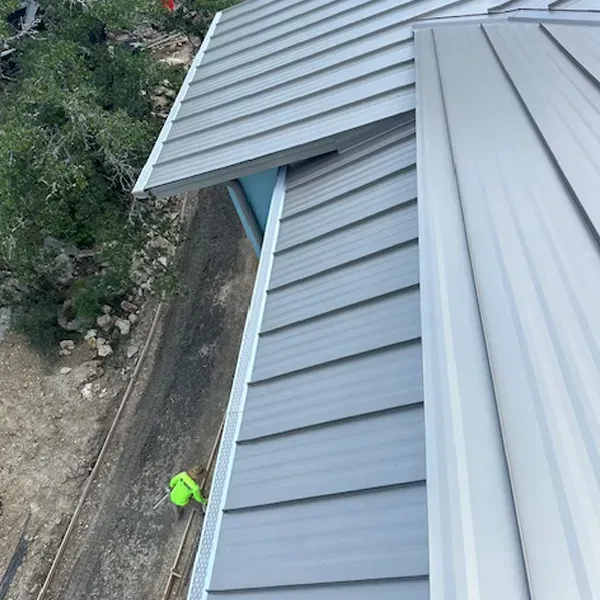Overhead view of a building's metal roof with gray siding and gutter against a backdrop of trees and a path.