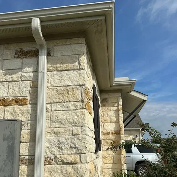 Corner of a stone house with white gutters against a blue sky, partially obscuring a white SUV.