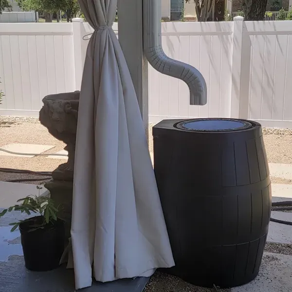 Rain barrel under a downspout, next to white fence. Beige curtain draped over a post.