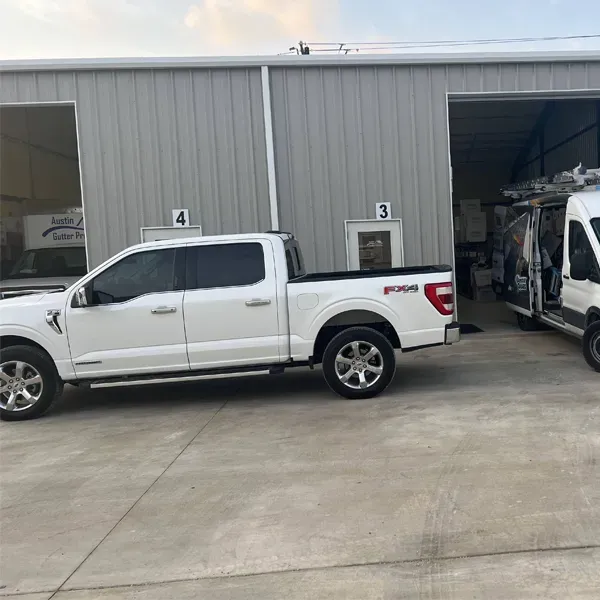 White pickup truck parked outside a gray industrial building with a van in an open bay door.