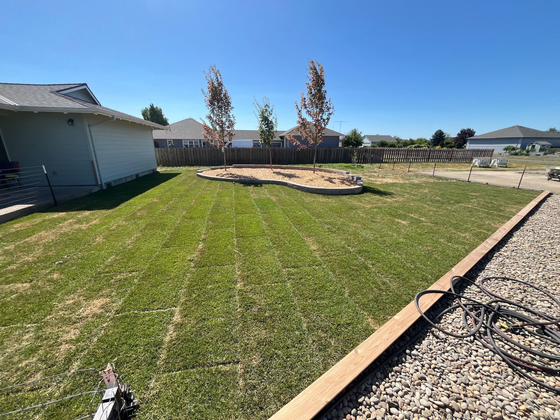 Well-manicured backyard with grass, trees, and a gravel border. Sunny day, house on the left, and neighborhood in the background.