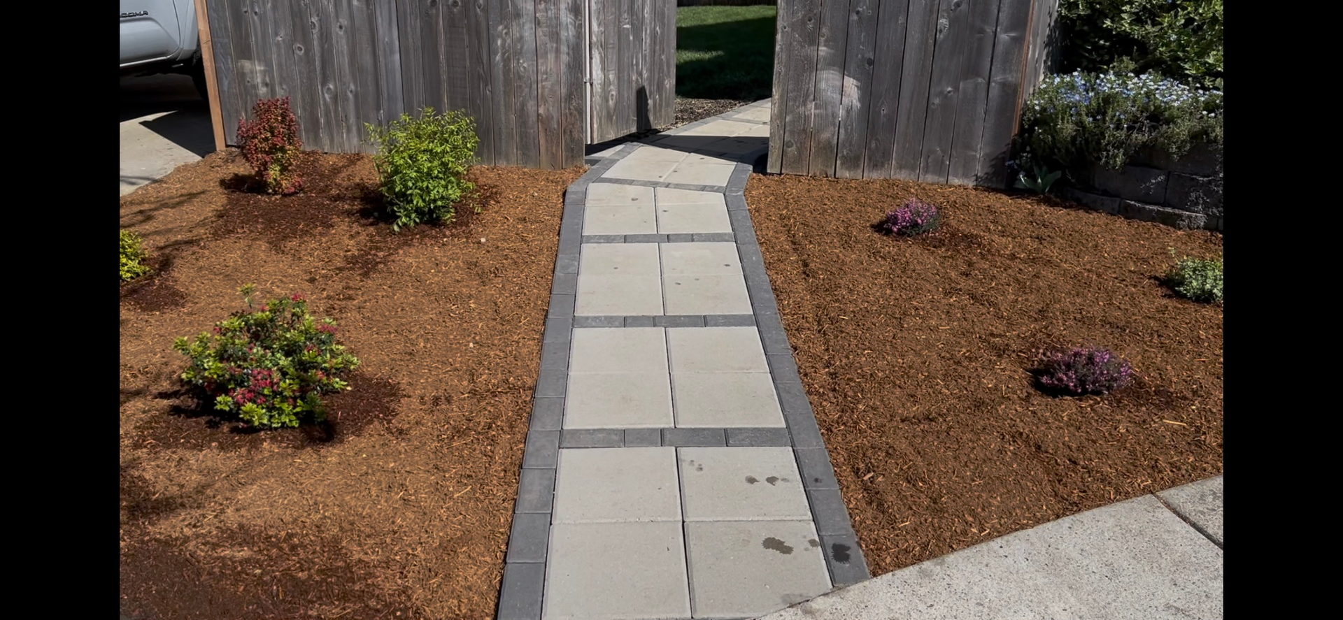 A garden path of square paving stones bordered by mulch and plants, leading to a wooden fence entrance.