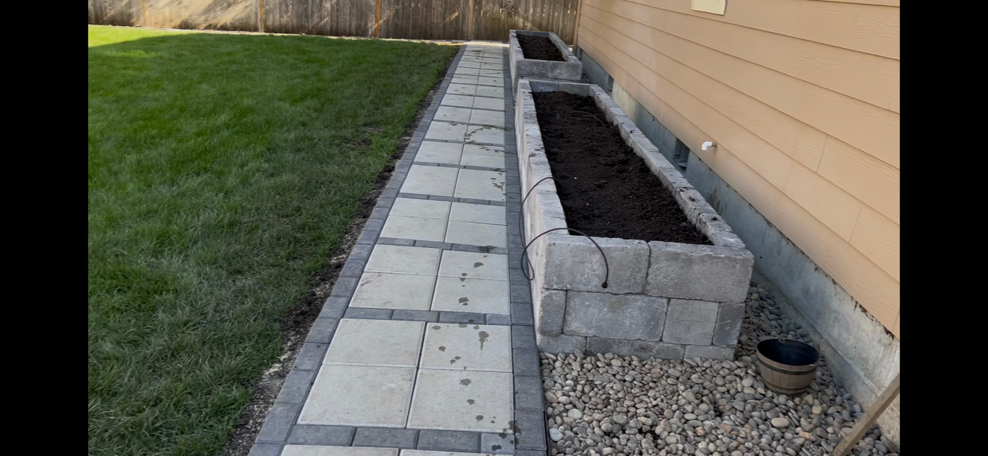 A backyard with a paved walkway and raised garden beds built with gray blocks next to a wooden house.