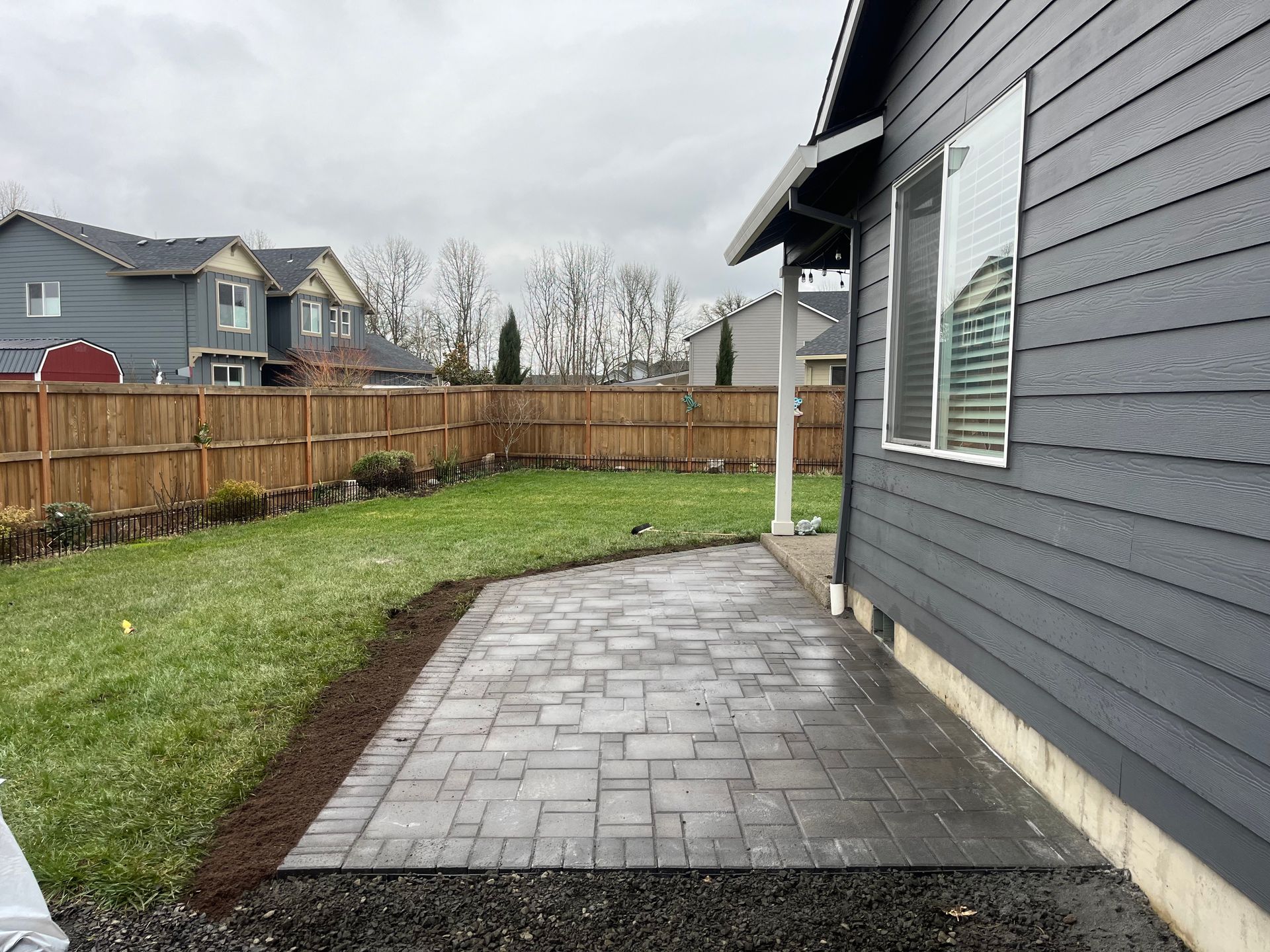 Backyard with a gray paver patio extending from a gray-sided house; green lawn, a wooden fence, and overcast sky are in the background.