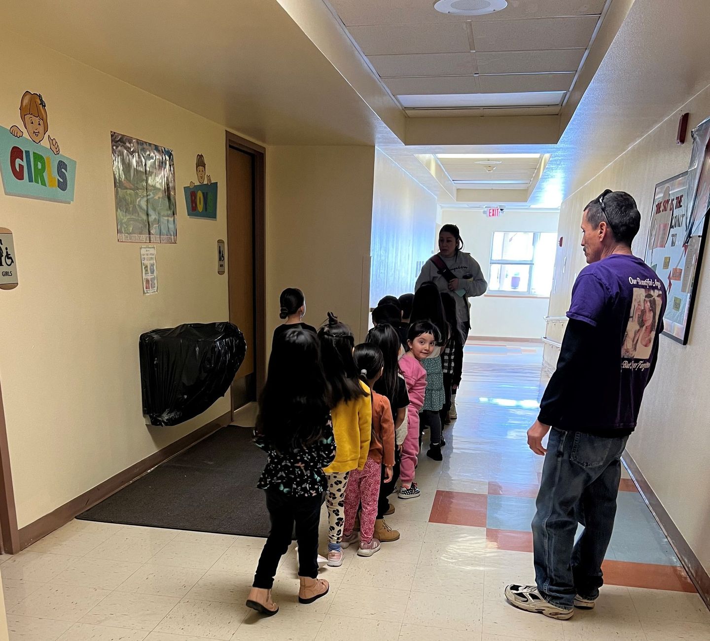 a man in a purple shirt is talking to a group of children in a hallway .