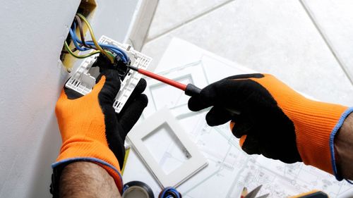 A person wearing orange gloves is working on an electrical outlet with a screwdriver.