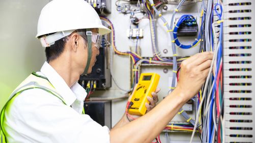 An electrician is working on an electrical box while holding a multimeter.