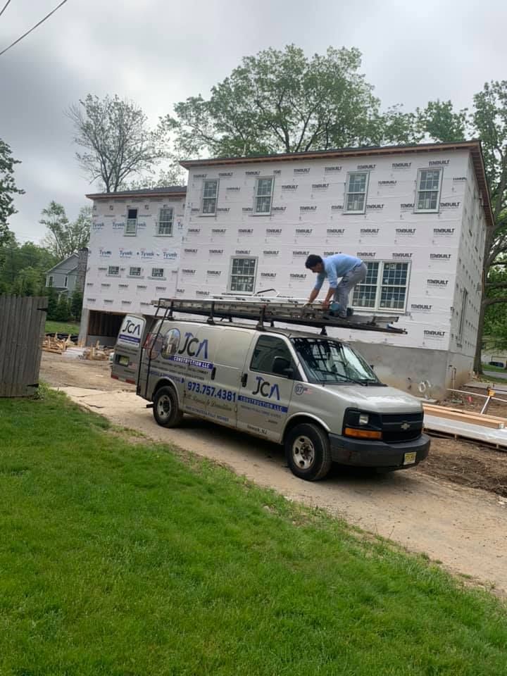 Van parked near a house under construction; worker on van roof. Gray van with company logo, partially finished house, cloudy sky.