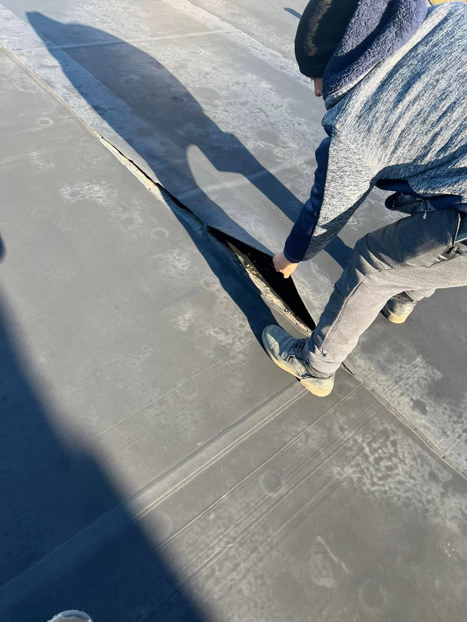 Person inspecting a tear in a dark, flat roof. The person is wearing gray clothing and boots.