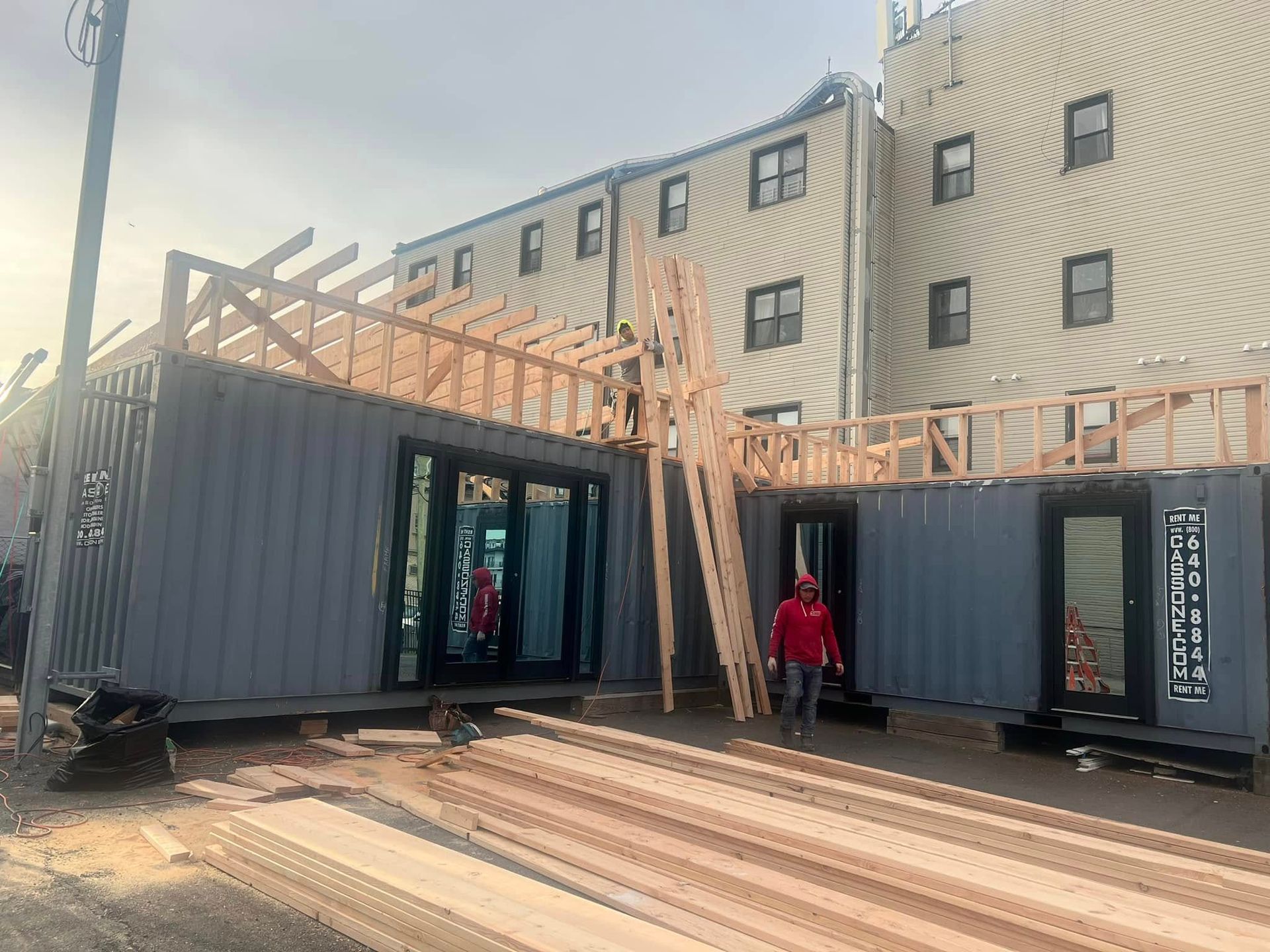 Construction site with shipping containers being converted into a building. A person in red walks by.