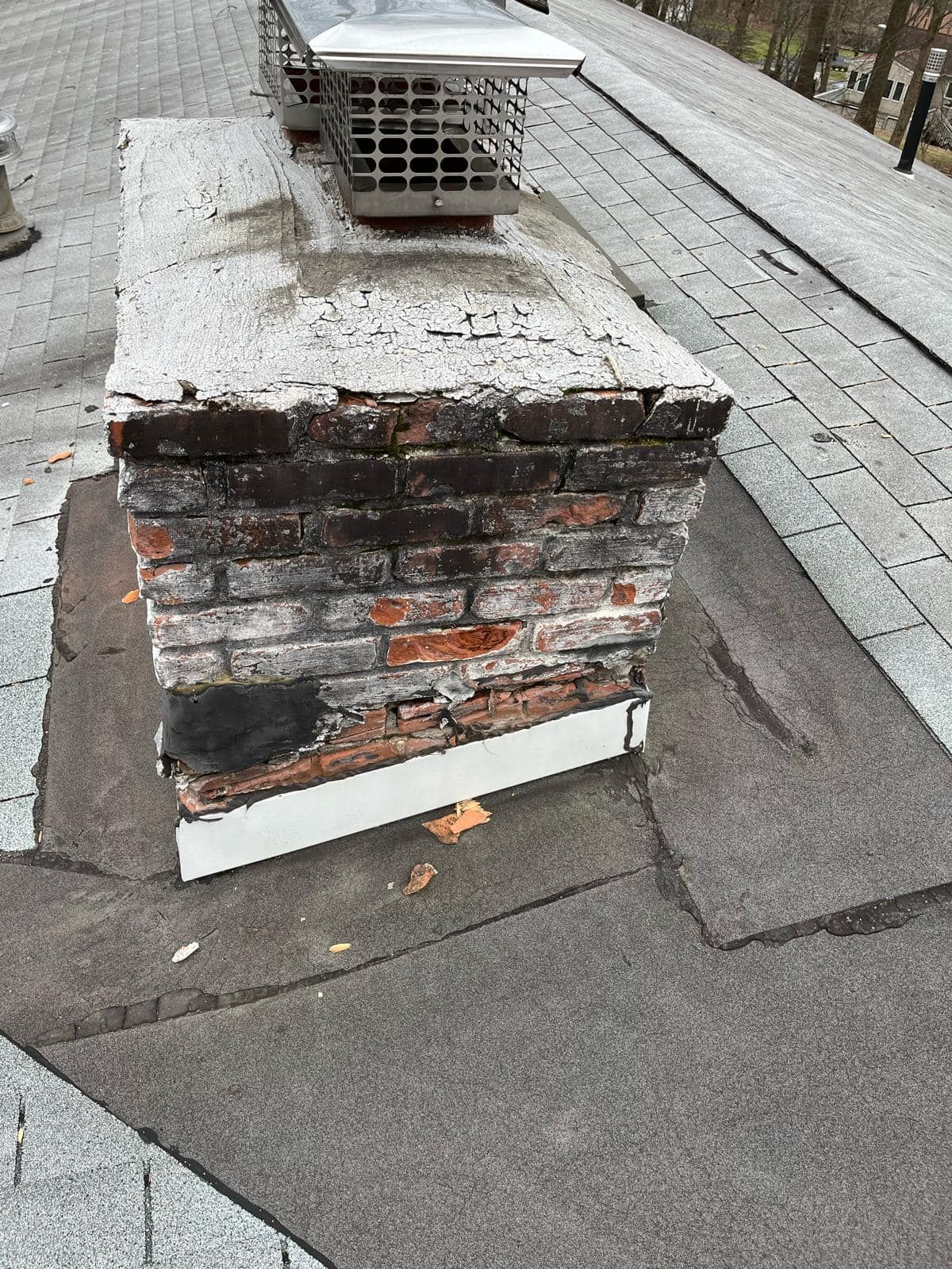 Brick chimney on a gray shingle roof, showing wear and tear, with a metal cap.