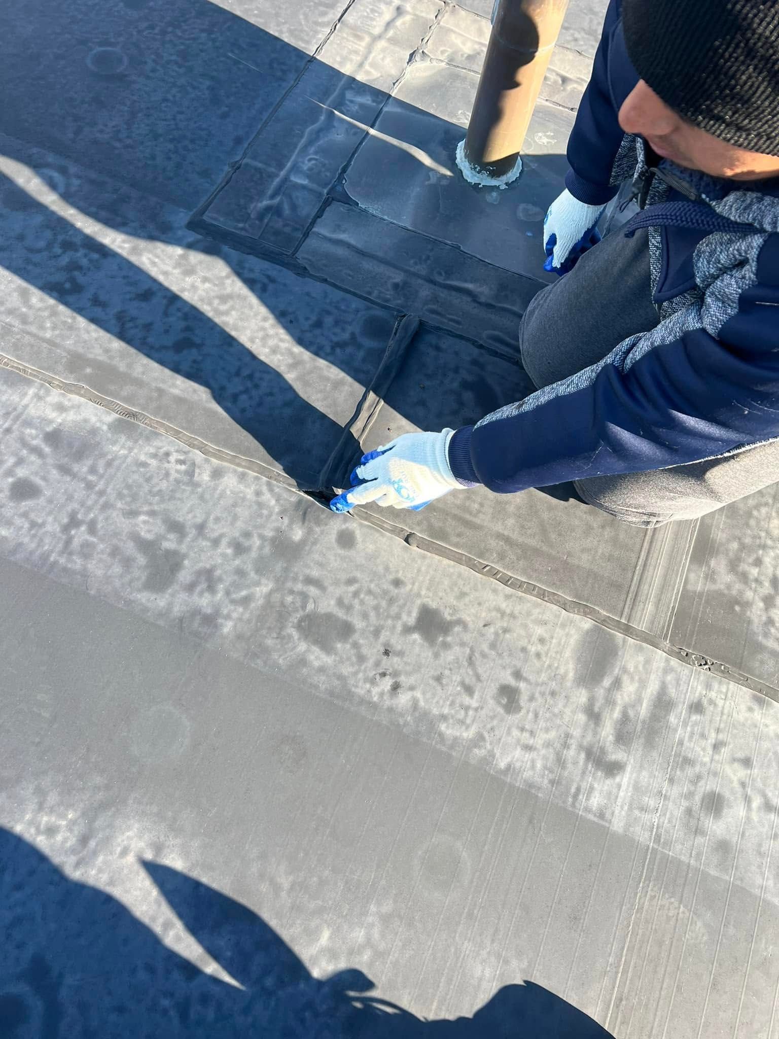 Person inspecting a dark roof, kneeling and wearing gloves. Sunlight casts shadows on the surface.