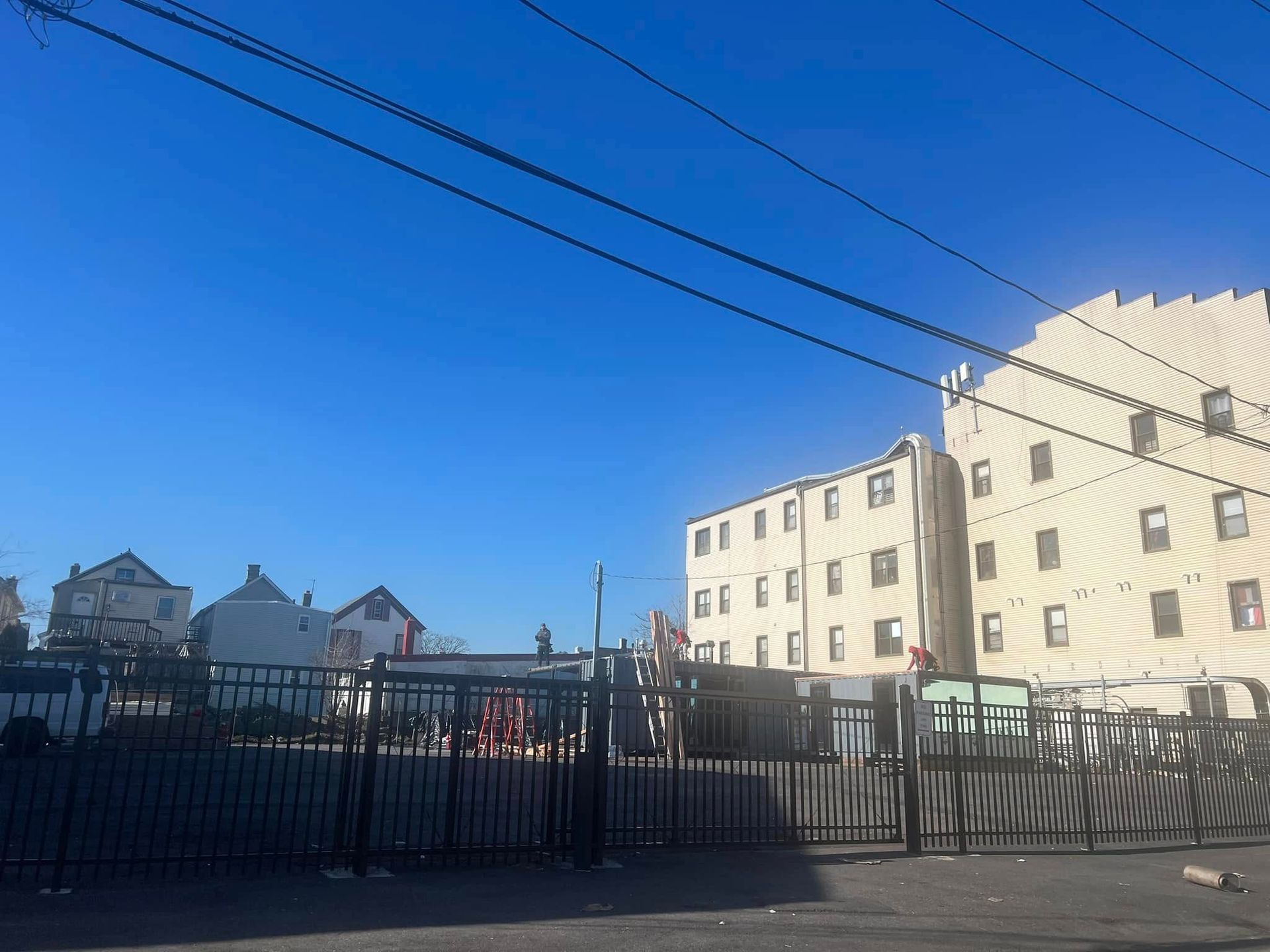 Black fence in front of an empty lot; apartment buildings and houses in the background under a blue sky.