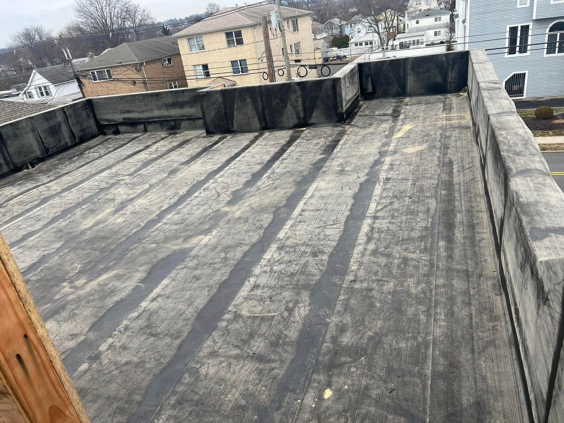 A flat, weathered roof with dark material and sealed seams, viewed from a high angle. Buildings in the background.