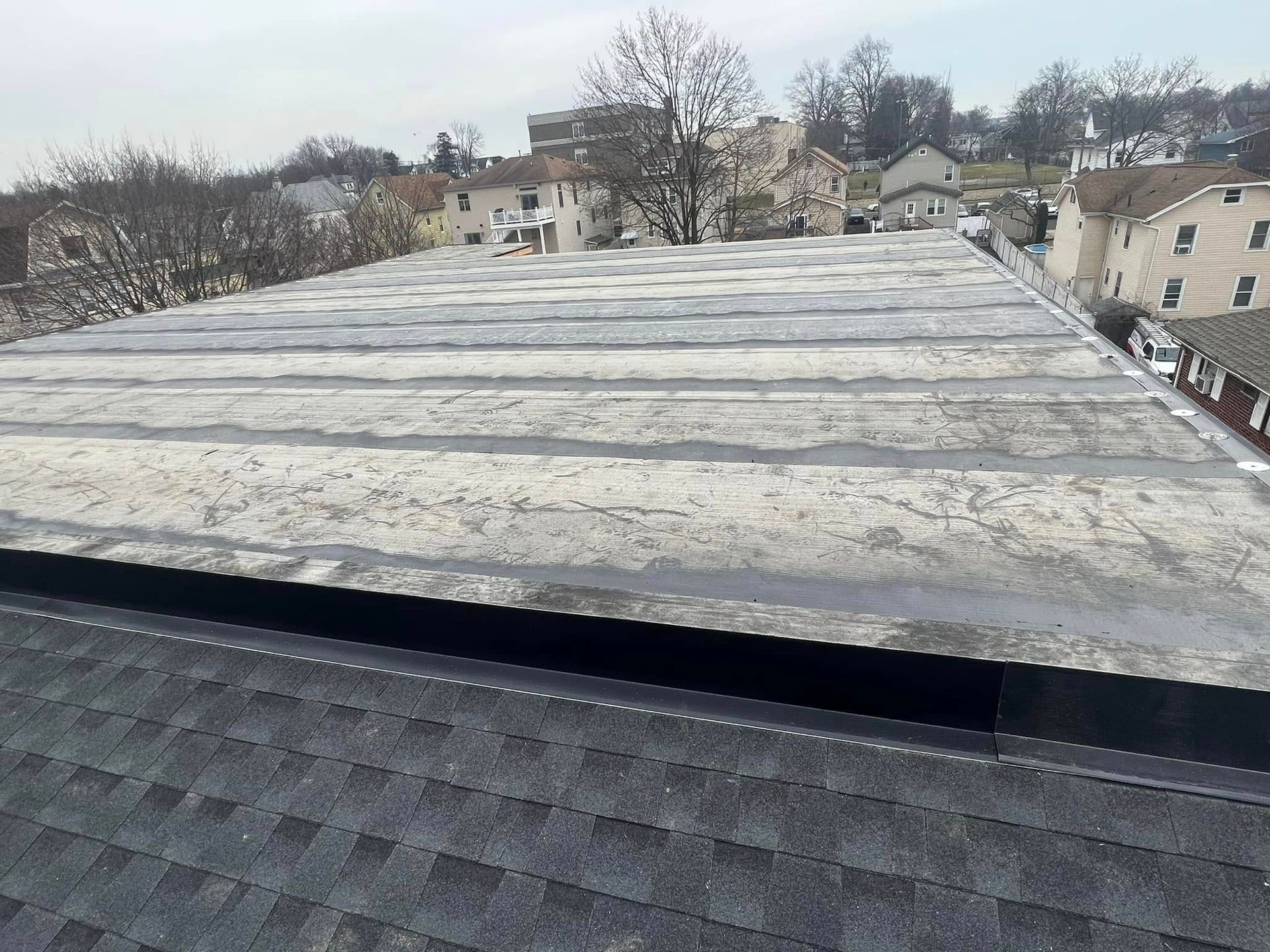 Gray flat roof with white patches, next to a black shingled roof, houses in the background.
