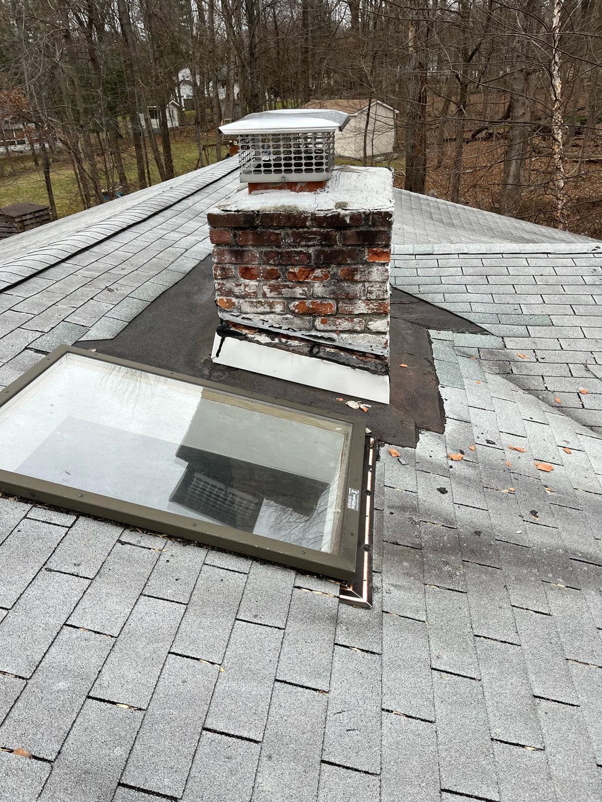 Chimney on a roof with a skylight, surrounded by gray asphalt shingles.