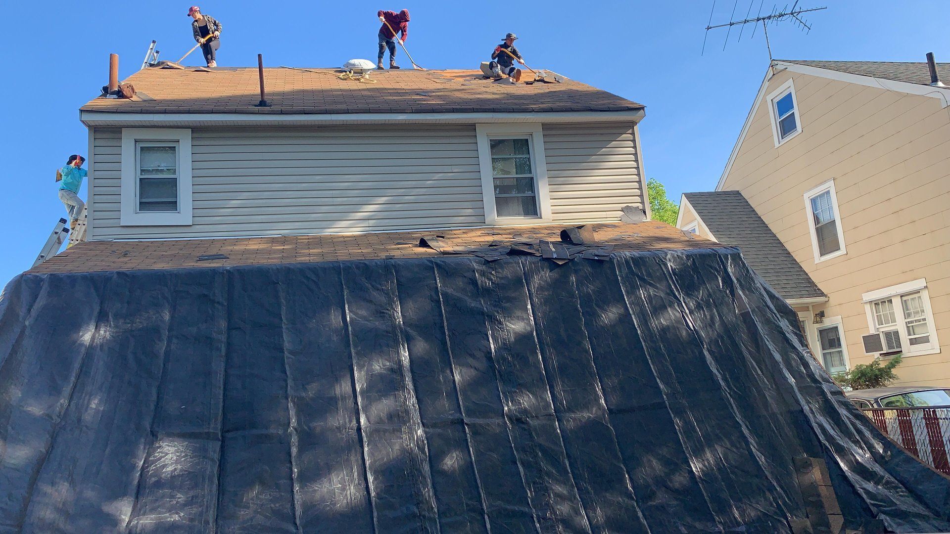 Roofers working on a house roof under a blue sky, covered by a tarp.