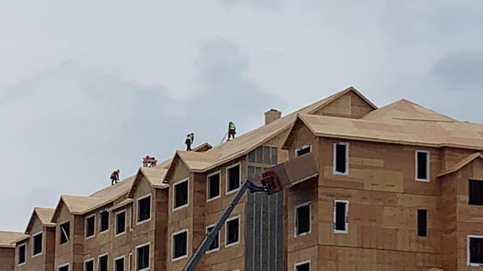 Construction workers on a multi-story building roof installing shingles. Gray sky background.