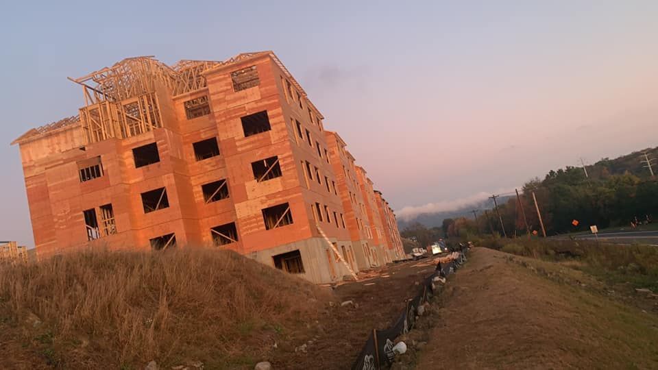 Brick building under construction on a hillside with surrounding vegetation and roadway.