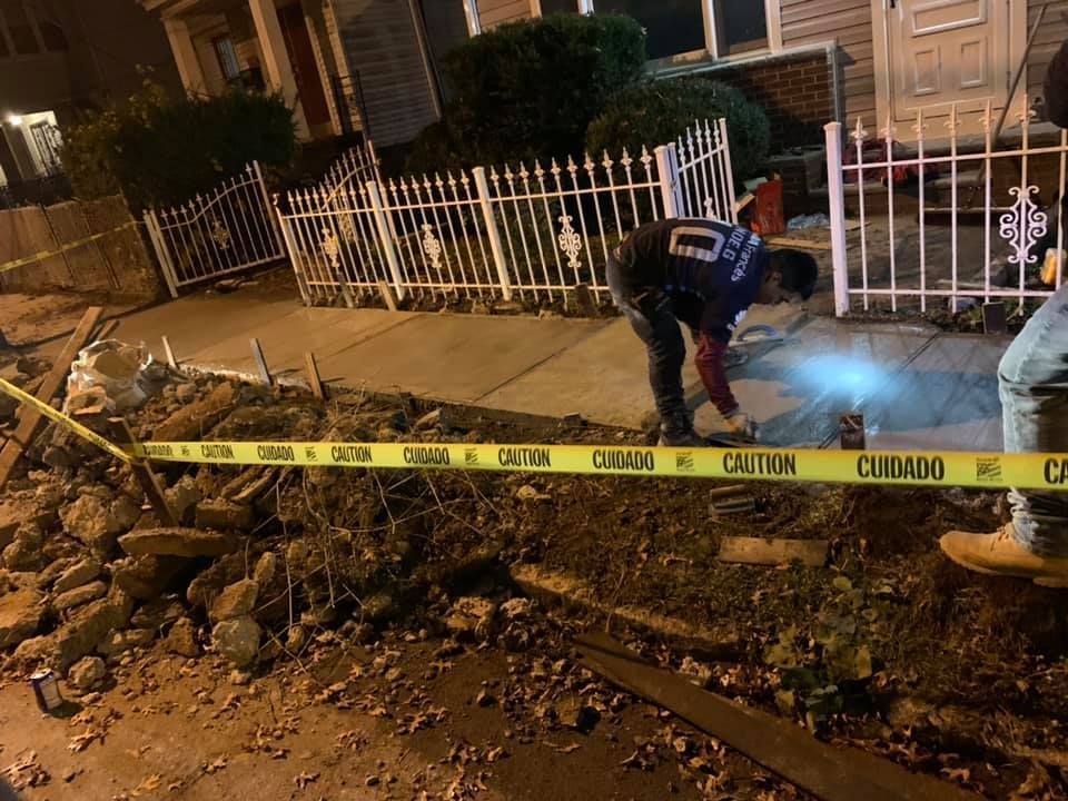 Police investigate a collapsed sidewalk near a house at night, yellow caution tape in foreground.