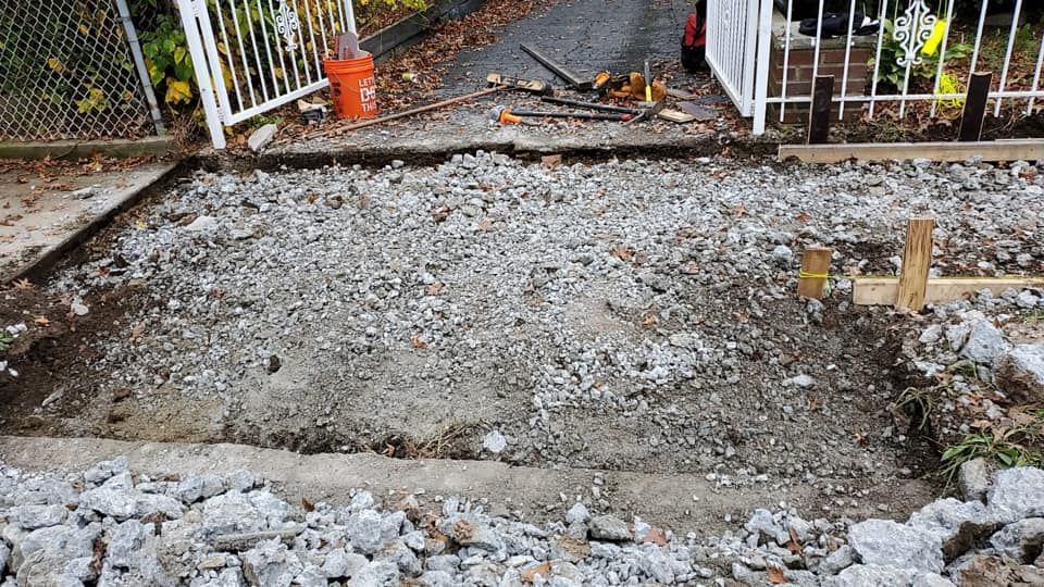 A driveway area with broken concrete, prepared for repaving, near a white gate.