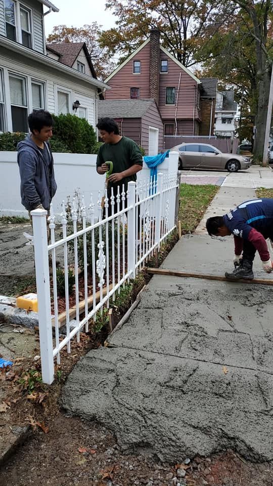 Three people working on a concrete sidewalk next to a white fence and houses.