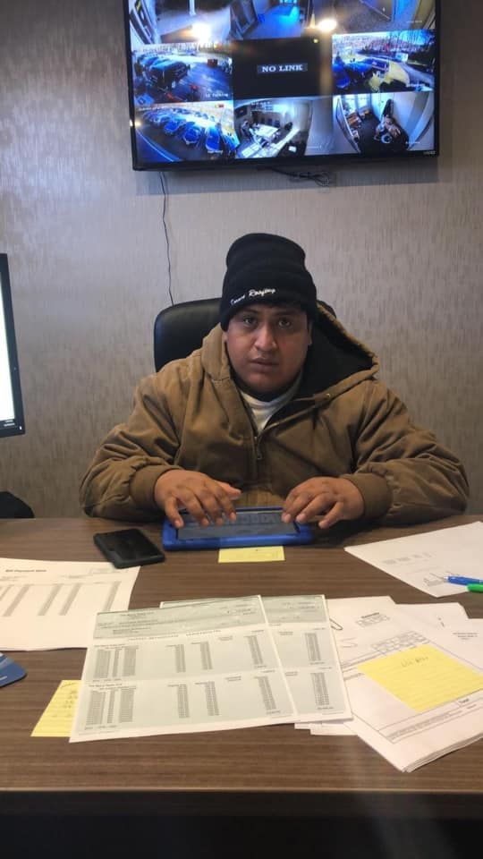 Person at desk wearing a beanie and jacket, looking at the camera. Monitors behind him display security footage. Papers and phone on desk.