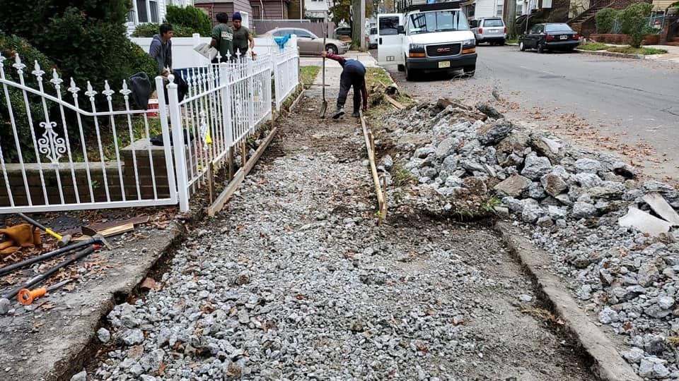 Workers repairing a sidewalk next to a white fence. Gray gravel, road, and parked vehicles.