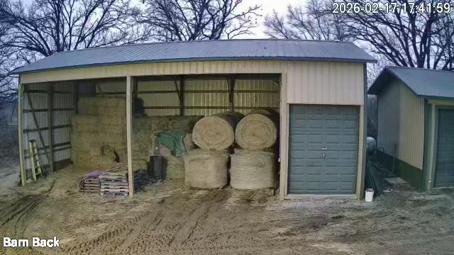 Open barn shed with hay bales and farm equipment inside, dirt ground outside, trees behind
