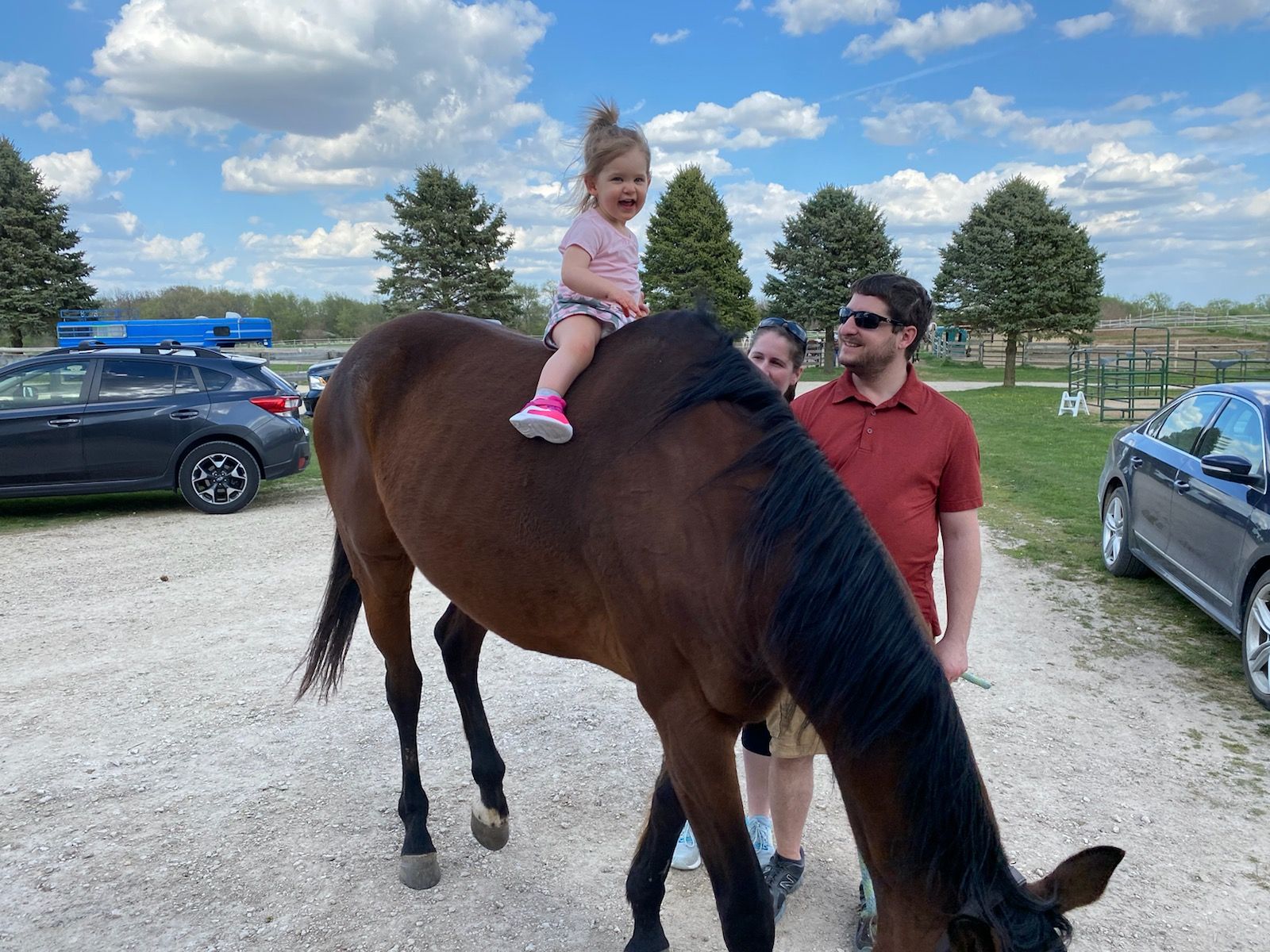 Child riding a brown horse while an adult leads it in a grassy parking area under a cloudy sky