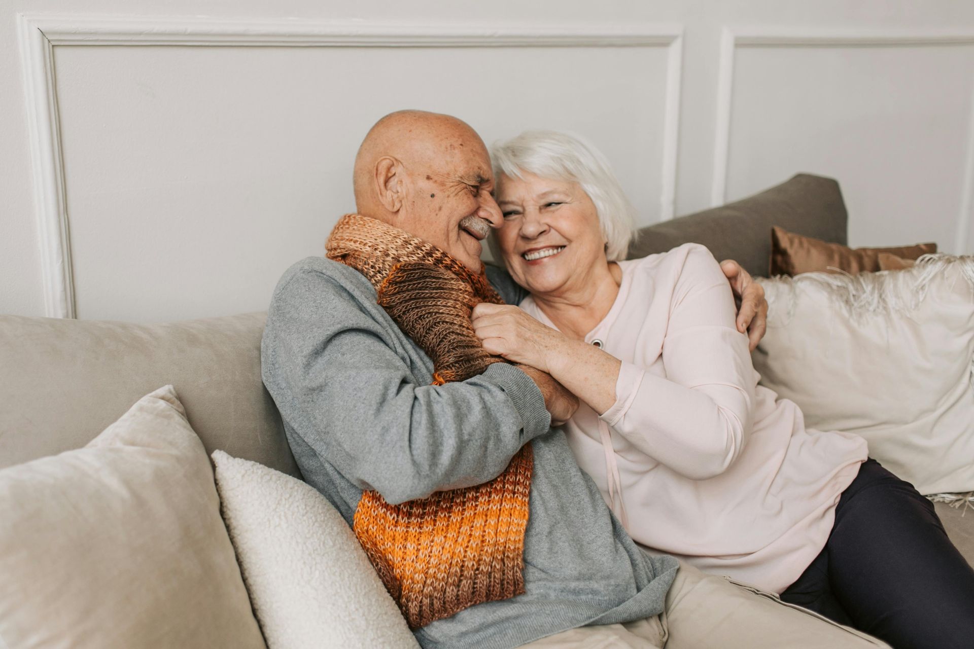 Couple de personnes âgées sur un canapé, souriant et enlacé. L'homme porte un foulard.