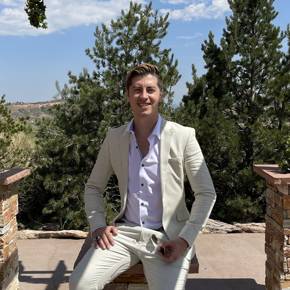 A man in a suit is sitting on a stone ledge with trees in the background