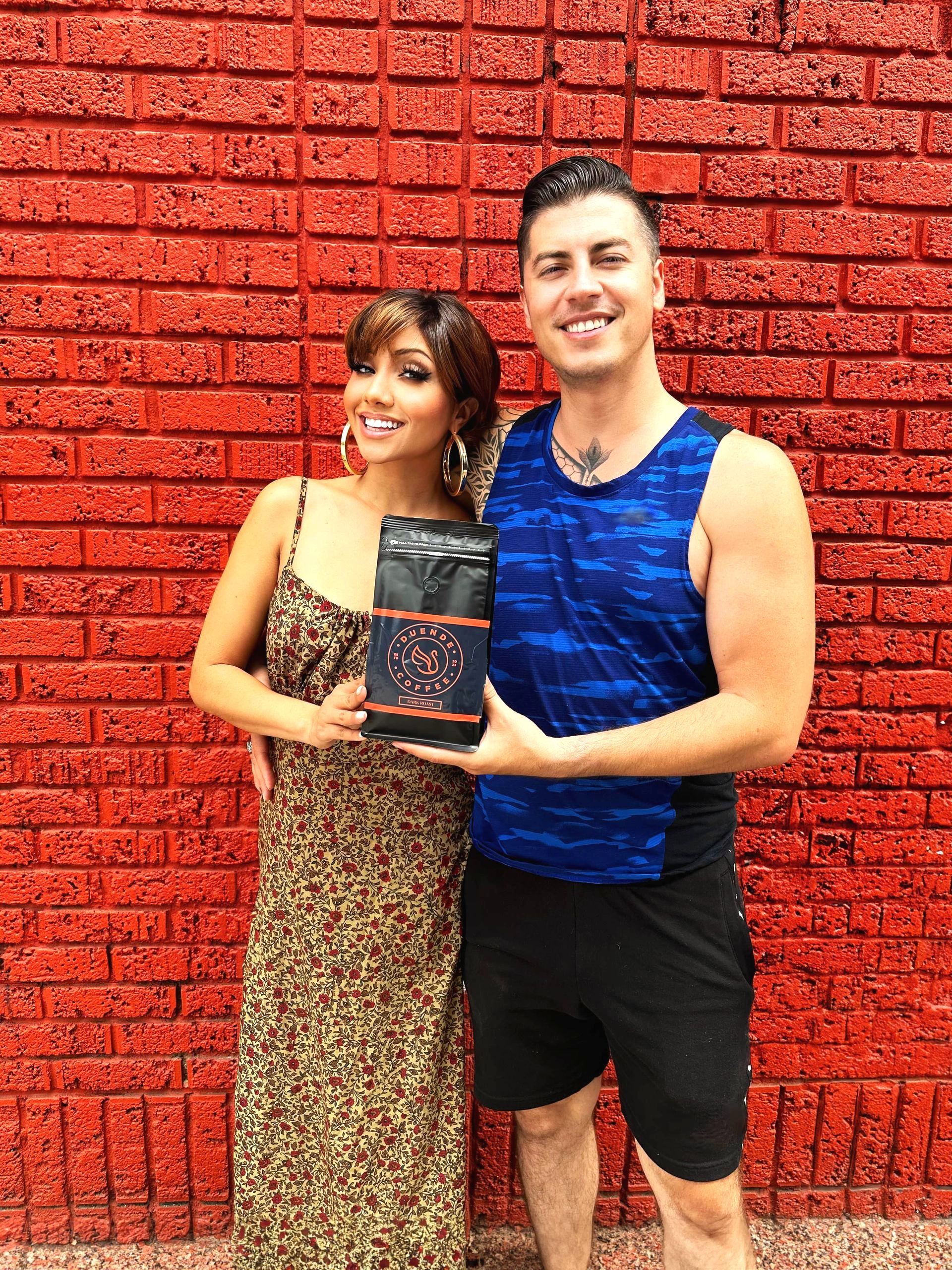 a man and a woman are standing in front of a red brick wall holding a bag of coffee .
