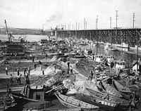 A black and white photo of boats in a harbor with a bridge in the background.