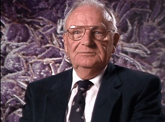 A man in a suit and tie stands in front of a purple background