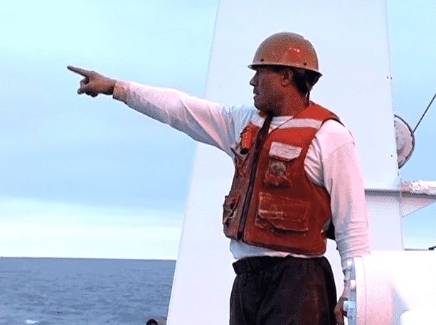 A man wearing a life vest and a hard hat points to the ocean