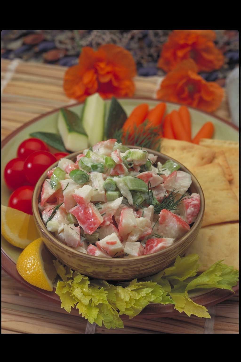 A bowl of crab salad is on a plate with crackers and vegetables.