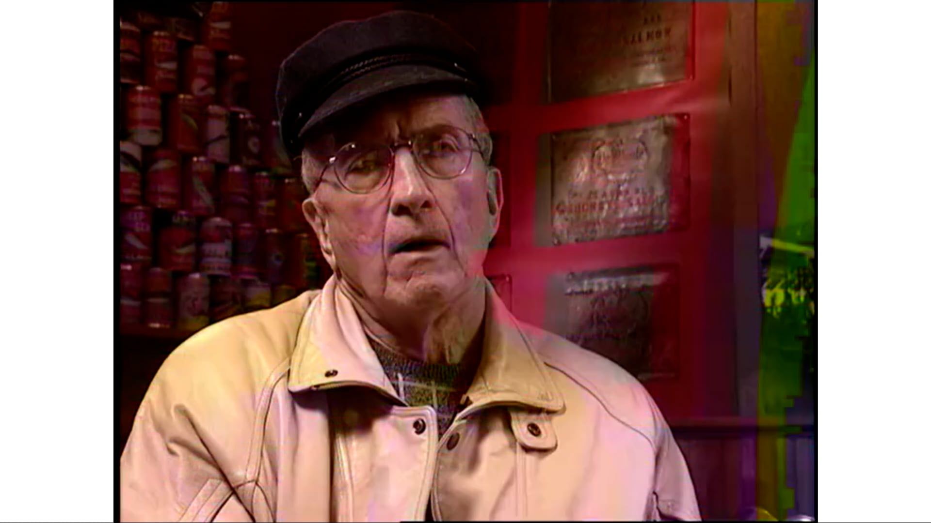 An elderly man wearing a hat and glasses is standing in front of a red wall.