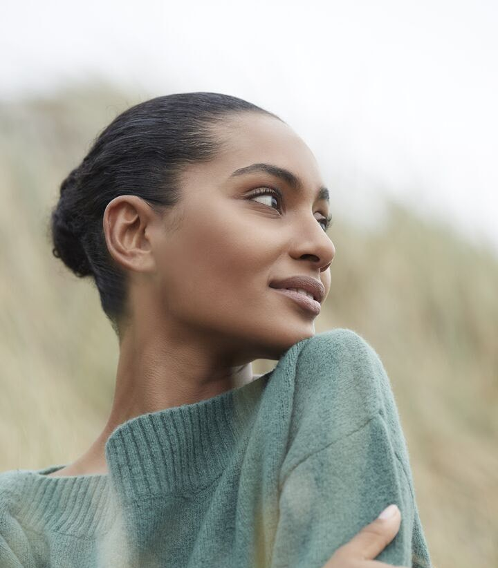 Femme en pull vert regardant hors champ, sur un fond extérieur flou.
