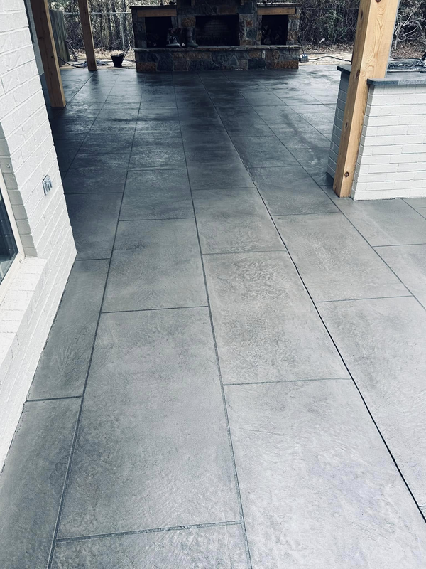 A textured gray concrete patio with stamped grid lines leads toward an outdoor stone fireplace under a covered porch.