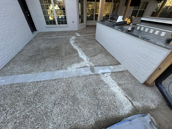 A concrete patio featuring repaired cracks with white filler, adjacent to a white brick outdoor kitchen structure.